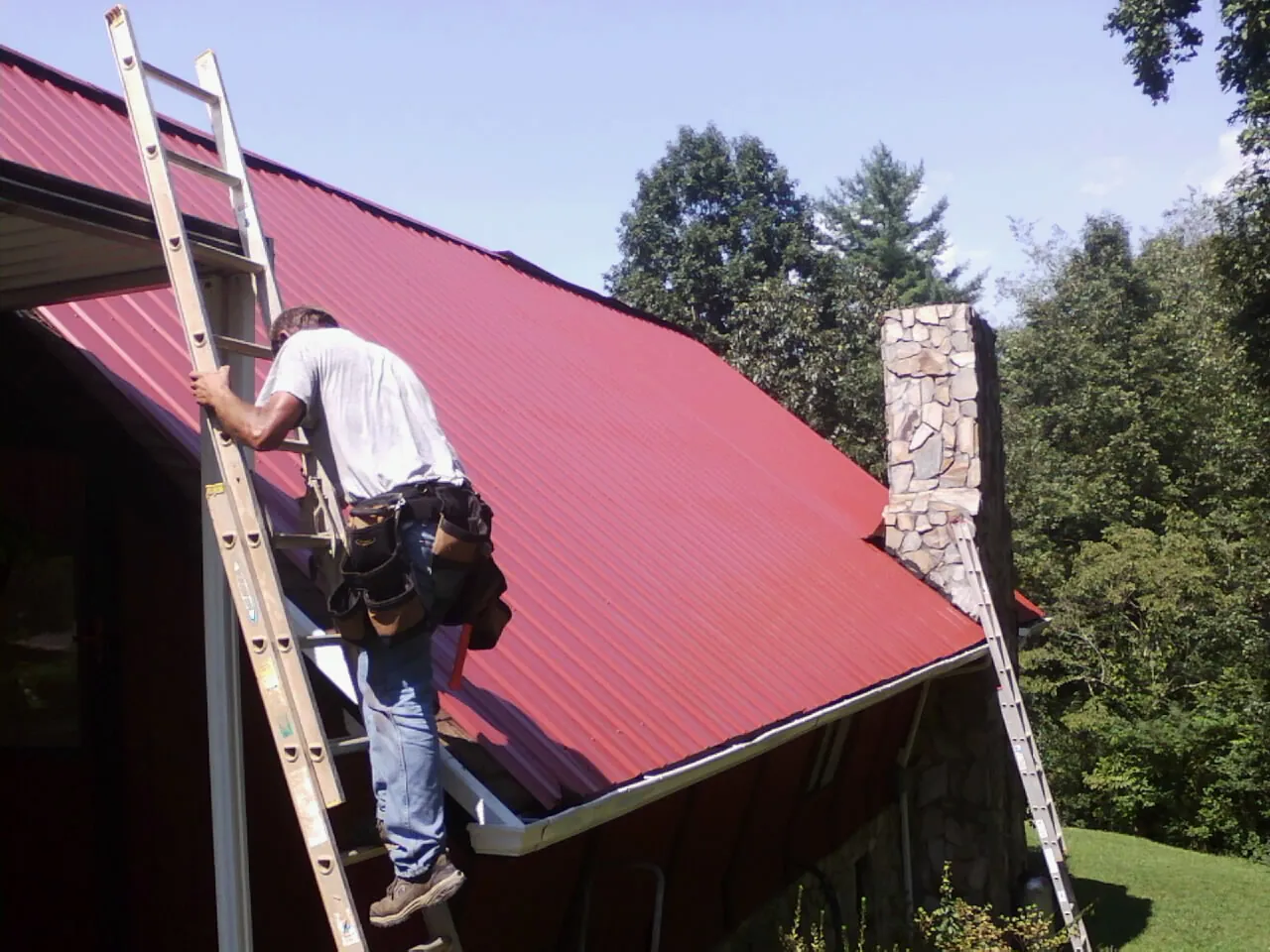 Roofer on a ladder beside a red metal roof and stone chimney — Soffit & Fascia Repair by Roofing Brothers in Farmland